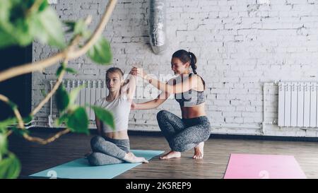 Die ziemlich blonde Yogaschülerin lernt Armdrehungen in der Haltung des Kuhgesichtes von einem freundlichen, fröhlichen Ausbilder beim persönlichen Training. Frauen sprechen, lächeln und genießen das Üben. Stockfoto