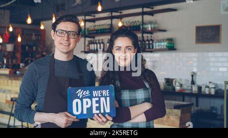 Fröhliche attraktive junge Menschen kleine Kaffeehausbesitzer halten wir sind offen Schild, während sie im modernen Café stehen. Eröffnung neuer Geschäfts- und Mitarbeiterkonzepte. Stockfoto