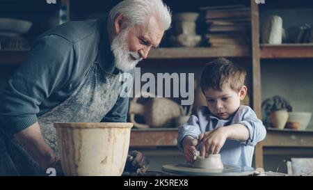 Konzentrierter Junge formt Ton in einen Keramiktopf auf dem drehenden Wurfrad und sein liebevoller erfahrener Großvater spricht mit ihm. Töpferei und Familientradition Konzept. Stockfoto