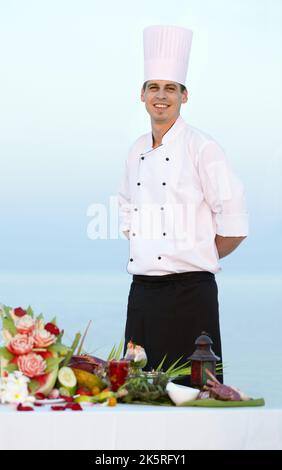 Ein kulinarisches Genie. Lächelnder junger Koch in Uniform, der stolz über sein Essen steht - Porträt. Stockfoto