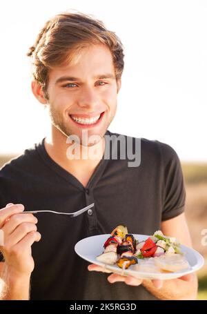 Wer nicht gerne im Sommer grillen kann. Porträt eines jungen Mannes, der sitzt und sein Essen genießt. Stockfoto