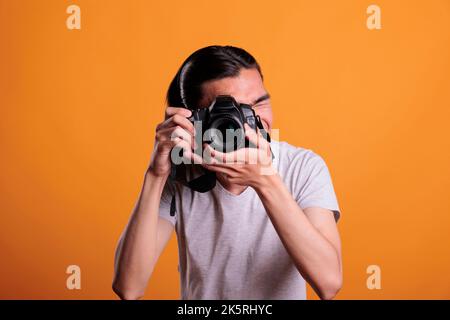 Fotograf, der Fotos mit einer professionellen dslr-Kamera macht, junger asiatischer Mann fotografiert, Vorderansicht, mittellange Aufnahme. Teenager, die im Sucher des Geräts suchen, kreatives Hobby, Fotoshooting Stockfoto