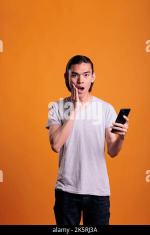 Schockierter asiatischer Mann beim Lesen von Nachrichten auf dem Smartphone, Online-Nachrichten, mitten in der Aufnahme. Verblüffter Teenager, der mit offenem Mund und Hand auf dem Gesicht stand, das Mobiltelefon hielt und über das Konzept des Klatschs schlauerte Stockfoto