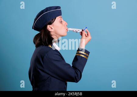 Stewardess trägt professionelle Luftfahrt Uniform, hält Flugzeugmodell, Flugbegleiter küssen Flugzeug Spielzeug. Air Hostess steht im Studio mit blauem Hintergrund, mittlere Aufnahme Stockfoto