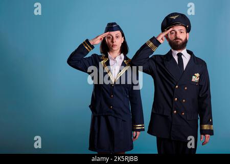 Flugzeugkapitän und Stewardess grüssen Porträt, Flugzeugbesatzung in professioneller Fluguniform. Selbstbewusster Flieger und Flughoster, Vorderansicht Studio mittellange Aufnahme auf blauem Hintergrund Stockfoto