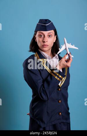 Flugbegleiter in der Luftfahrt Uniform hält Flugzeugmodell, professionelle Stewardess spielt mit Flugzeug. Lufthostess, die auf kommerzielles Flugzeugspielzeug schaut, Seitenansicht Studio mittlere Aufnahme Stockfoto