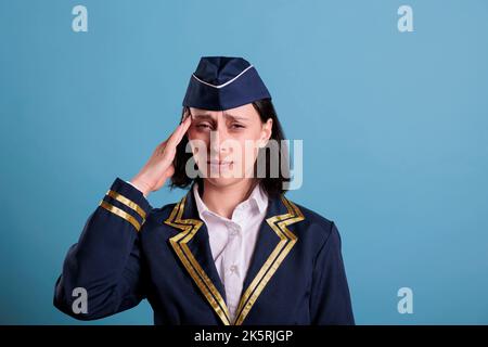 Flugbegleiter mit Migränesymptom, berühren Tempel mit Fingern, Blick auf die Kamera mit Grimasse. Unglückliche Stewardess in Uniform, die unter Kopfschmerzen leidet und sich unwohl fühlt Stockfoto