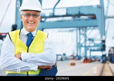 Glücklich, hier zu sein. Porträt eines Hafenarbeiters, der inmitten der Schifffahrt am Hafen steht. Stockfoto