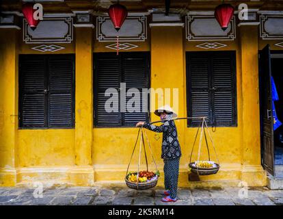 Eine weibliche Obstverkäuferin steht vor einem alten gelben Gebäude in der Altstadt von Hoi an, Vietnam. Stockfoto