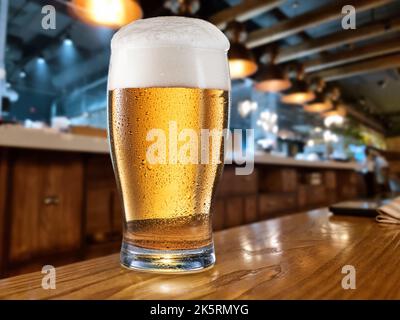 Gekühltes Glas Bier auf dem Holztisch. Unscharfer Barinnenraum im Hintergrund. Stockfoto