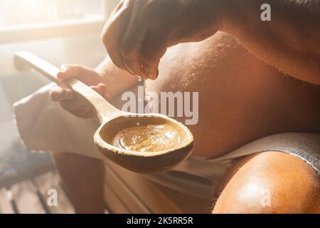 Alter Mann tropft Wasser in einen alten Holzlöffel in der finnischen Sauna, Spa und warmen Temperatur Bad Therapie Konzept Bild. Stockfoto