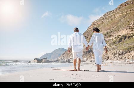 Rückansicht eines Seniorenpaares mit gemischtem Rennen, das einen romantischen Spaziergang am Strand Unternehmen und sich an einem sonnigen Sommertag im Freien die Hände halten kann Stockfoto