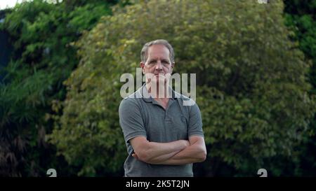 Ernsthafter Mann mittleren Alters, der im Park steht und die Kamera anschaut. Porträt eines älteren Mannes mit gekreuzten Armen Stockfoto