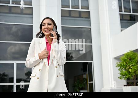 Eine glückliche und attraktive junge asiatische Geschäftsfrau oder Managerin unterhält sich gerne mit ihrer Freundin am Telefon, während sie aus dem Firmengebäude läuft Stockfoto