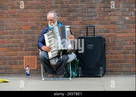 LONDON - 21. Mai 2022: Älterer Straßenmusiker mit weißem Bart spielt Akkordeon neben alter roter Ziegelmauer Stockfoto