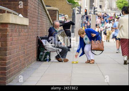 LONDON - 21. Mai 2022: Frau gibt älteren Straßenmusikern mit weißem Bart und Akkordeon Geld. Touristen im Hintergrund Stockfoto