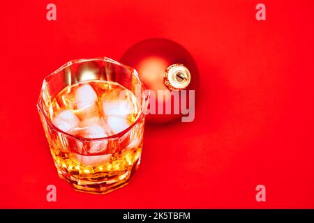 Glas Whiskey mit Eiswürfeln und weihnachtskugel auf rotem Hintergrund - weihnachtsgetränk-Konzept Stockfoto