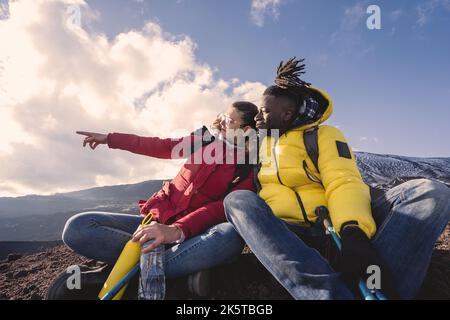 Multiethnisches Paar junger Menschen, die sich auf dem Gipfel des Berges entspannen, auf dem Boden sitzen und mit den Fingern wegschauen - Wanderlust Wanderer gucken Stockfoto