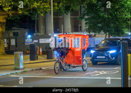 LONDON - 21. Mai 2022: Der Rikscha-Fahrer trägt im nächtlichen Verkehr Gesichtsmaske und Mütze Stockfoto