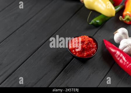 Ajika auf schwarzem Hintergrund. Scharfe Chilipfeffer-Harissa-Sauce. Hausgemachte Harissa in einer Schüssel. Kopierbereich, Draufsicht, Ebene Ebene. Stockfoto