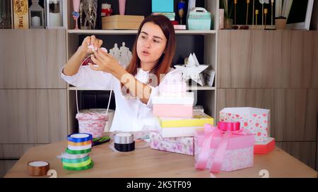 Geschenkverpackung. Attraktive Frau, Designer von Geschenken und Dekorationen zeigt Training online, zeichnet Videoblog, Webinar, sitzt vor dem Smartphone, Kamera. Online-Verkauf. Hochwertige Fotos Stockfoto
