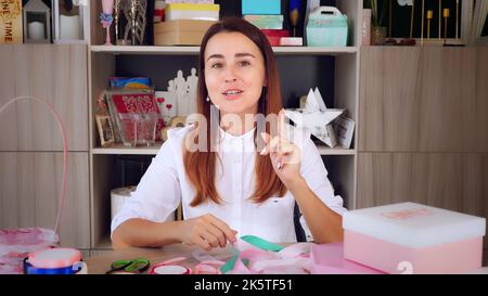 Geschenkverpackung. Attraktive Frau, Designer von Geschenken und Dekorationen zeigt Training online, zeichnet Videoblog, Webinar, sitzt vor dem Smartphone, Kamera. Online-Verkauf. Hochwertige Fotos Stockfoto