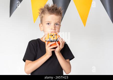 Halloween Kinder Junge mit Kürbis Eimer essen Süßigkeiten auf einem weißen Studio Hintergrund mit einer Girlande. Bereit für den Urlaub. Stockfoto