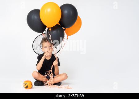 Ein fröhlicher, fröhlicher Junge in einem Zaubererhut mit einem Kürbis-förmigen Eimer und schwarzen und orangefarbenen Ballons, der auf einem weißen Studiohintergrund sitzt. Bereit für Stockfoto