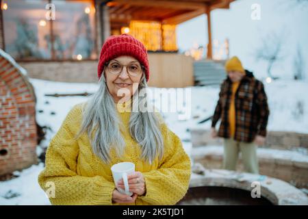 Glückliche ältere Frau mit einer Tasse heißen Tee, die im Winter vor dem Haus steht. Stockfoto