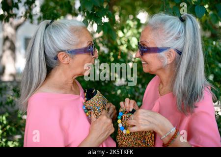 Ältere Frauen Zwillinge in farbenfroher Kleidung mit außergewöhnlichen Handtaschen in der Stadt, reden und lächeln. Stockfoto