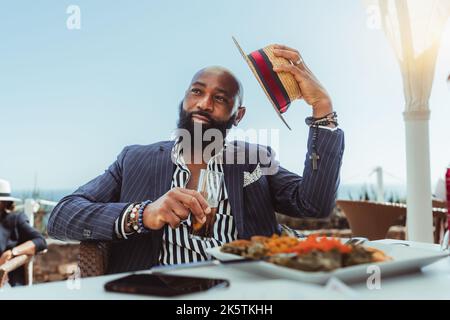 Porträt eines stämchenhaften, kahlen Afrikaners mit schwarzem Bart und in einem maßgeschneiderten Anzug, der in einem Küstenstraßenrestaurant mit einem Glas b zu Mittag gegessen hat Stockfoto