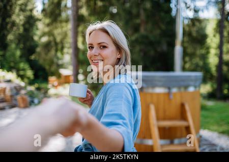 Glückliche junge Frau, die die Hände hält, aus persönlicher Perspektive betrachtet, mit einer Tasse Kaffee vor der Badewanne im Freien stehend. Stockfoto