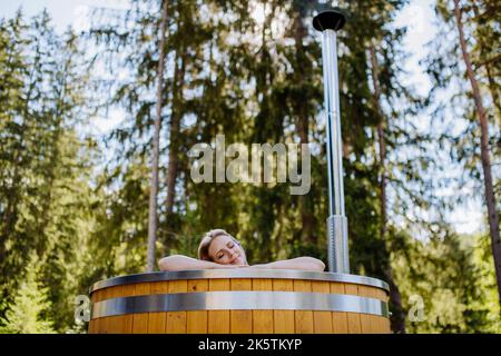 Junge Frau, die in der Holzbadewanne mit Kamin Holz verbrennt und Wasser im Hinterhof in den Bergen erwärmt. Stockfoto