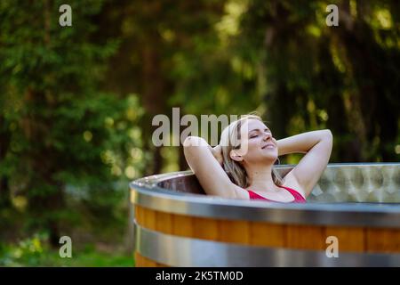 Junge Frau, die in der Holzbadewanne mit Kamin Holz verbrennt und Wasser im Hinterhof in den Bergen erwärmt. Stockfoto