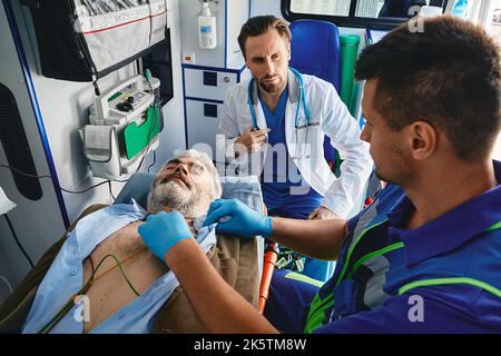 Herzpatient im Krankenwagen. Sanitäter, die EKG an einem männlichen Vizitum durchführen, der auf einer Bahre liegt, wobei die Elektroden auf seiner Brust im Krankenwagen platziert wurden Stockfoto