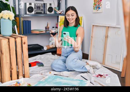 Junge hispanische Künstlerin, die im Kunststudio mit einem Smartphone Wein trinkt Stockfoto