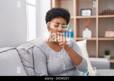 Junge afroamerikanische Frau trägt Virtual-Reality-Brille auf dem Sofa sitzen Gefühl unwohl und Husten als Symptom für Kälte oder Bronchitis. Gesundheit Stockfoto