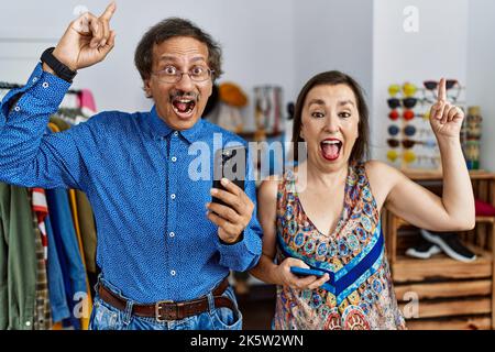 Interrassisches Paar im mittleren Alter im Einzelhandelsgeschäft mit Smartphone lächelnd und überrascht und mit Fingern und erhobenen Armen nach oben zeigend. Stockfoto
