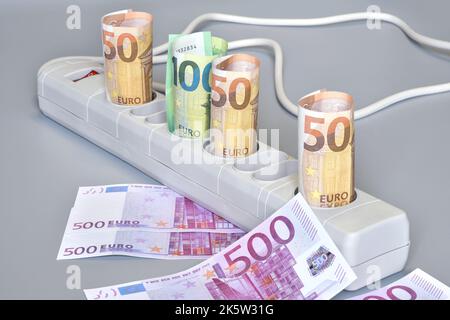 Euro-Banknoten gerollt und in die Steckdosen einer elektrischen Steckdosenleiste auf grauem Hintergrund eingeführt. Konzept für die steigenden Kosten Stockfoto