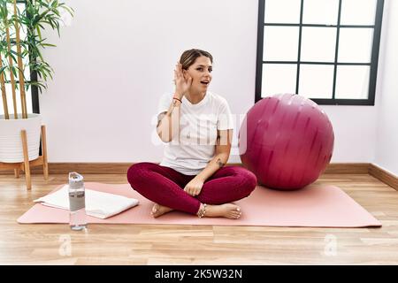 Junge hispanische Frau, die im Sportzentrum trainiert, lächelt mit der Hand über dem Ohr und hört Gerüchten oder Klatsch zu. Taubheitskonzept. Stockfoto