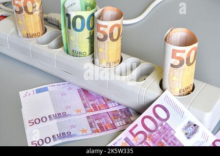 Euro-Banknoten gerollt und in die Steckdosen einer elektrischen Steckdosenleiste auf grauem Hintergrund eingeführt. Konzept für die steigenden Kosten Stockfoto
