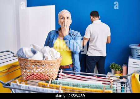 Hispanische Mutter und Sohn hängen Kleidung an Wäscheleine, die den Mund mit der Hand bedeckt, schockiert und Angst vor Fehlern. Überraschter Ausdruck Stockfoto