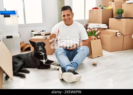 Junger latein, der mit Hund taka away Sushi isst, sitzt auf dem Boden in einem neuen Zuhause. Stockfoto