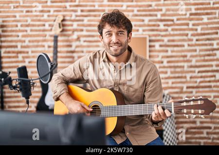 Junger Musiker, der im Musikstudio lächelnd und selbstbewusst klassische Gitarre spielt Stockfoto