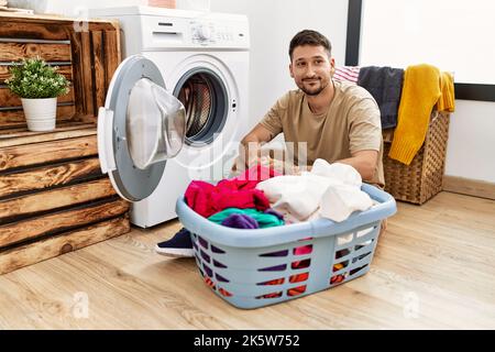 Ein junger, gutaussehender Mann, der schmutzige Wäsche in die Waschmaschine legt und lächelnd zur Seite schaut und denkend wegstarrt. Stockfoto