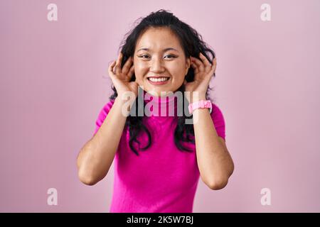 Junge asiatische Frau über rosa Hintergrund lächelnd ziehen Ohren mit Fingern, lustige Geste. Audition Problem Stockfoto