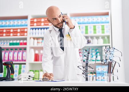 Junger kahlköpfiger Apotheker, der auf dem Smartphone spricht und auf einem Dokument in der Apotheke schreibt Stockfoto