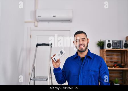 Der hispanische Reparaturmann, der mit der Klimaanlage arbeitet, sieht positiv aus und ist glücklich, steht und lächelt mit einem selbstbewussten Lächeln, das die Zähne zeigt Stockfoto