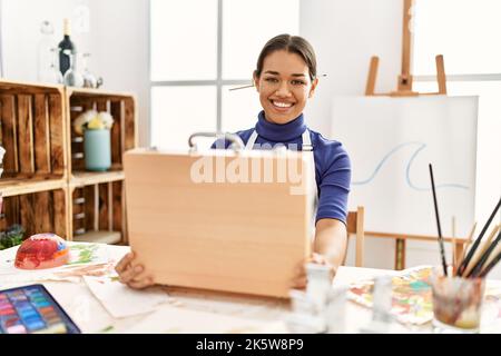 Junge latinerin lächelt selbstbewusst und eröffnet eine Aktentasche im Kunststudio Stockfoto