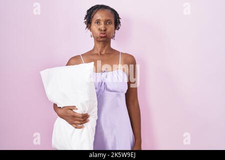 Afrikanische Frau mit Dreadlocks tragen Pyjama umarmt Kissen puffing Wangen mit lustigen Gesicht. Mund mit Luft aufgeblasen, verrückten Ausdruck. Stockfoto